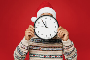 Wall Mural - Merry young man wear Christmas sweater Santa hat posing hold show covering hiding face with clock time five to twelve isolated on plain red background. Happy New Year 2023 celebration holiday concept.