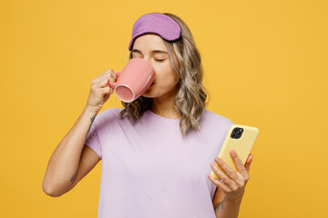 Wall Mural - Calm young woman wears purple pyjamas jam sleep eye mask rest relax at home hold use mobile cell phone cup drink tea isolated on plain yellow background studio portrait. Good mood night nap concept.