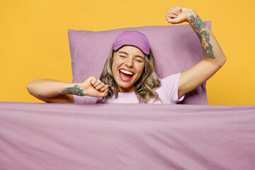 Wall Mural - Fun young woman she wears purple pyjamas jam sleep eye mask rest relax at home lies wrap covered under blanket duvet stretch hands yawn isolated on plain yellow background Good mood night nap concept.