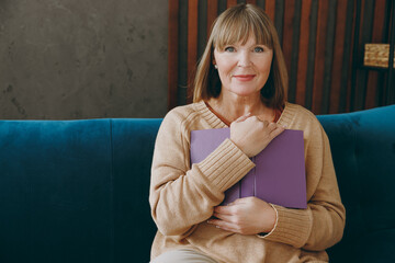 Smiling happy elderly woman 50s year old wearing casual clothes glasses sit on blue sofa read book novel look camera stay at home flat rest relax spend free spare time in living room indoor grey wall