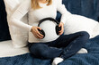 Young pretty pregnant woman holds black headphones on belly sitting on bed, prenatal music listening. Happy childbearing period, caring for unborn baby. Female wears white long sleeve, blue jeans