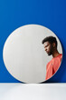 © Clique Images - Vertical creative studio shot of handsome young Black man wearing coral sweater in round mirror reflection, blue background