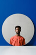 © Clique Images - Vertical creative studio shot of stylish young Black man wearing coral sweater looking at camera, round mirror reflection, blue background