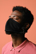 © Clique Images - Vertical close-up conceptual portrait of unrecognizable stylish young African American man wearing black fabric mask on face posing on camera with eyes closed against peach pink background