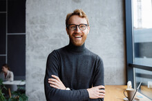 A Young Red-haired Guy, A Programmer Or An Entrepreneur In Glasses, In A Stylish Cafe Looks At The Camera And Smiles. Freelancer Works Remotely. Online Communication. Small Business.