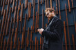 © YURII MASLAK - A young red-haired guy, a businessman in glasses, against the backdrop of urban style, checks mail on a smartphone, communicates in a messenger and drinks coffee. Online communication.