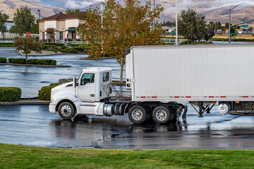 Day cab white big rig semi truck delivering food cargo in refrigerator semi trailer to local customer standing on the parking lot early morning