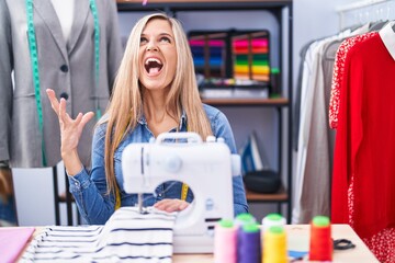 Sticker - Blonde woman dressmaker designer using sew machine crazy and mad shouting and yelling with aggressive expression and arms raised. frustration concept.