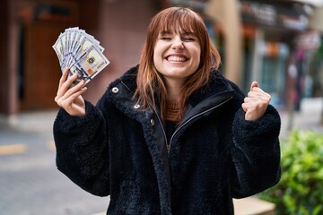 Canvas Print - Young beautiful woman holding dollars banknotes screaming proud, celebrating victory and success very excited with raised arm