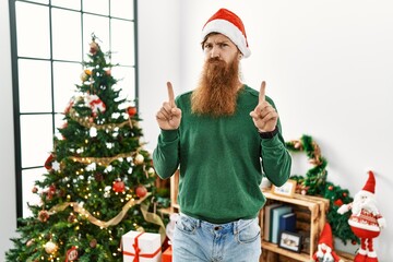 Sticker - Redhead man with long beard wearing christmas hat by christmas tree pointing up looking sad and upset, indicating direction with fingers, unhappy and depressed.