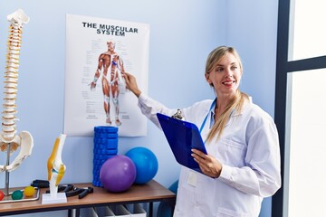 Wall Mural - Young blonde woman wearing physiotherpist uniform pointing to anatomy paper at physiotherapy clinic