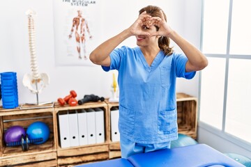 Wall Mural - Middle age hispanic physiotherapist woman working at pain recovery clinic doing heart shape with hand and fingers smiling looking through sign