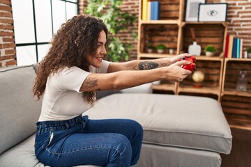 Wall Mural - Young beautiful hispanic woman playing video game sitting on sofa at home