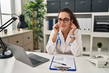 Poster - Young hispanic woman wearing doctor uniform and stethoscope covering ears with fingers with annoyed expression for the noise of loud music. deaf concept.