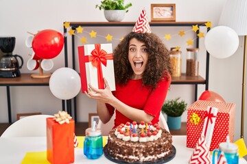 Poster - Hispanic woman with curly hair celebrating birthday with cake and present celebrating crazy and amazed for success with open eyes screaming excited.