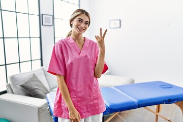 Wall Mural - Young blonde woman working as physiotherapist at home showing and pointing up with fingers number two while smiling confident and happy.