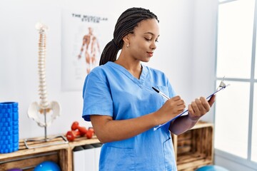 Wall Mural - Young african american woman wearing physio therapist uniform writing on clipboard at clinic