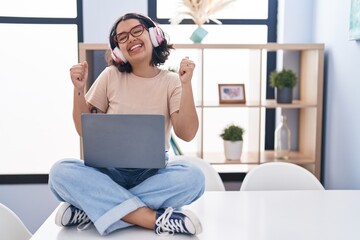 Poster - Young hispanic woman using laptop sitting on the table wearing headphones very happy and excited doing winner gesture with arms raised, smiling and screaming for success. celebration concept.