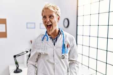 Canvas Print - Young blond man wearing doctor uniform and stethoscope at clinic angry and mad screaming frustrated and furious, shouting with anger. rage and aggressive concept.