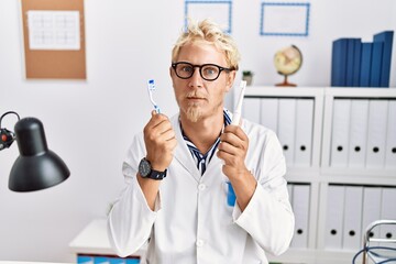 Canvas Print - Young blond man working at dentist clinic holding electric toothbrush and teethbrush skeptic and nervous, frowning upset because of problem. negative person.