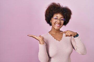 Poster - Young african american woman standing over pink background amazed and smiling to the camera while presenting with hand and pointing with finger.