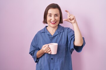 Sticker - Middle age hispanic woman drinking a cup coffee smiling and confident gesturing with hand doing small size sign with fingers looking and the camera. measure concept.