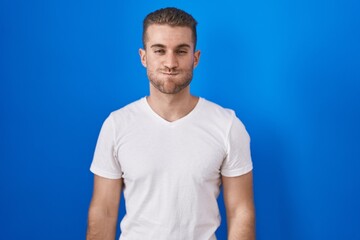 Canvas Print - Young caucasian man standing over blue background puffing cheeks with funny face. mouth inflated with air, crazy expression.