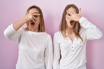 Sticker - Middle age mother and young daughter standing over pink background peeking in shock covering face and eyes with hand, looking through fingers with embarrassed expression.