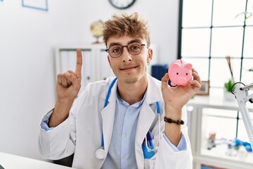 Wall Mural - Young caucasian doctor man wearing doctor uniform holding piggy bank at the clinic smiling with an idea or question pointing finger with happy face, number one