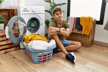 Wall Mural - Young caucasian man putting dirty laundry into washing machine smiling looking to the side and staring away thinking.