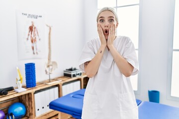 Canvas Print - Young caucasian woman working at pain recovery clinic afraid and shocked, surprise and amazed expression with hands on face