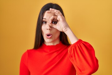 Canvas Print - Young hispanic woman standing over yellow background doing ok gesture shocked with surprised face, eye looking through fingers. unbelieving expression.