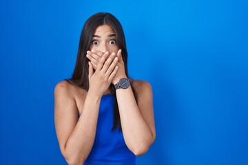 Sticker - Hispanic woman standing over blue background shocked covering mouth with hands for mistake. secret concept.