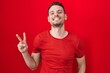 © Krakenimages.com - Young hispanic man standing over red background smiling looking to the camera showing fingers doing victory sign. number two.