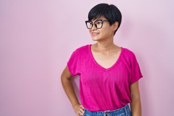 Wall Mural - Young asian woman with short hair standing over pink background looking away to side with smile on face, natural expression. laughing confident.