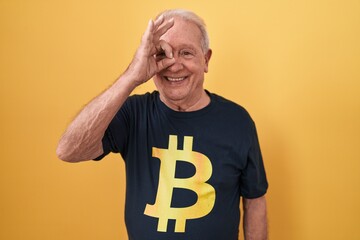 Canvas Print - Senior man with grey hair wearing bitcoin t shirt doing ok gesture with hand smiling, eye looking through fingers with happy face.