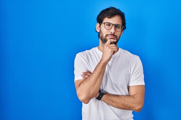 Poster - Handsome latin man standing over blue background looking confident at the camera smiling with crossed arms and hand raised on chin. thinking positive.