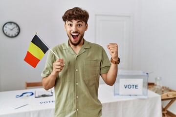 Sticker - Young arab man at political campaign election holding belgium flag screaming proud, celebrating victory and success very excited with raised arms