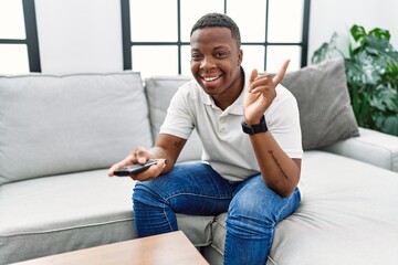 Poster - Young african man watching tv holding television remote control smiling happy pointing with hand and finger to the side