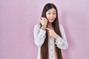 Poster - Chinese young woman standing over pink background in hurry pointing to watch time, impatience, looking at the camera with relaxed expression