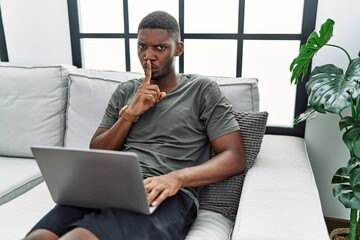 Poster - Young african american man using laptop at home sitting on the sofa asking to be quiet with finger on lips. silence and secret concept.