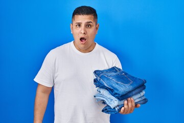 Canvas Print - Hispanic young man holding stack of folded jeans scared and amazed with open mouth for surprise, disbelief face