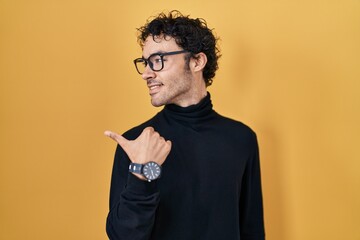 Wall Mural - Hispanic man standing over yellow background smiling with happy face looking and pointing to the side with thumb up.