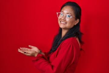 Poster - Asian young woman standing over red background pointing aside with hands open palms showing copy space, presenting advertisement smiling excited happy