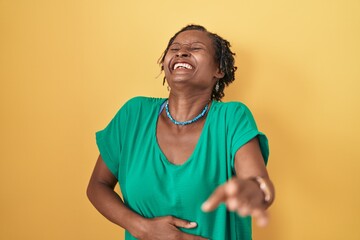 African woman with dreadlocks standing over yellow background laughing at you, pointing finger to the camera with hand over body, shame expression