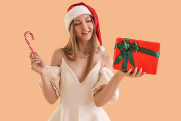 Poster - Young woman in Santa hat with Christmas gift and candy cane on beige background