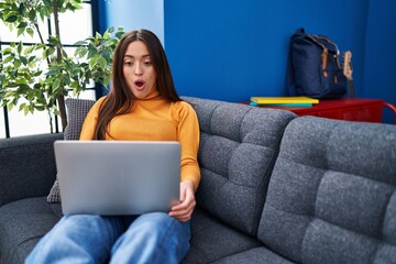 Sticker - Young brunette woman working using computer laptop sitting on the sofa scared and amazed with open mouth for surprise, disbelief face