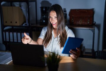 Poster - Young brunette woman working at the office at night approving doing positive gesture with hand, thumbs up smiling and happy for success. winner gesture.