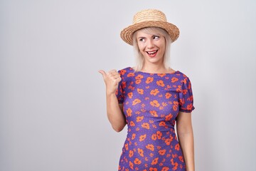 Poster - Young caucasian woman wearing flowers dress and summer hat smiling with happy face looking and pointing to the side with thumb up.