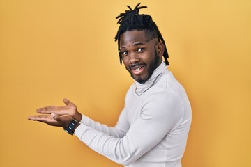 Wall Mural - African man with dreadlocks wearing turtleneck sweater over yellow background pointing aside with hands open palms showing copy space, presenting advertisement smiling excited happy